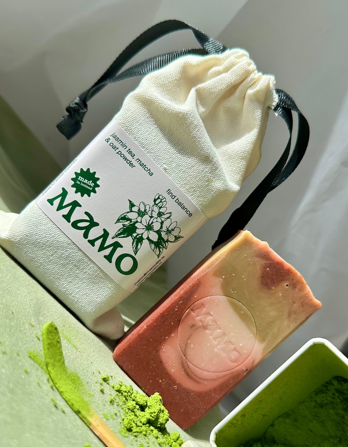 A handmade soap bar with pink and cream layers, displayed next to a white drawstring pouch labeled 'MAMO' and a green powder container on a light green surface.