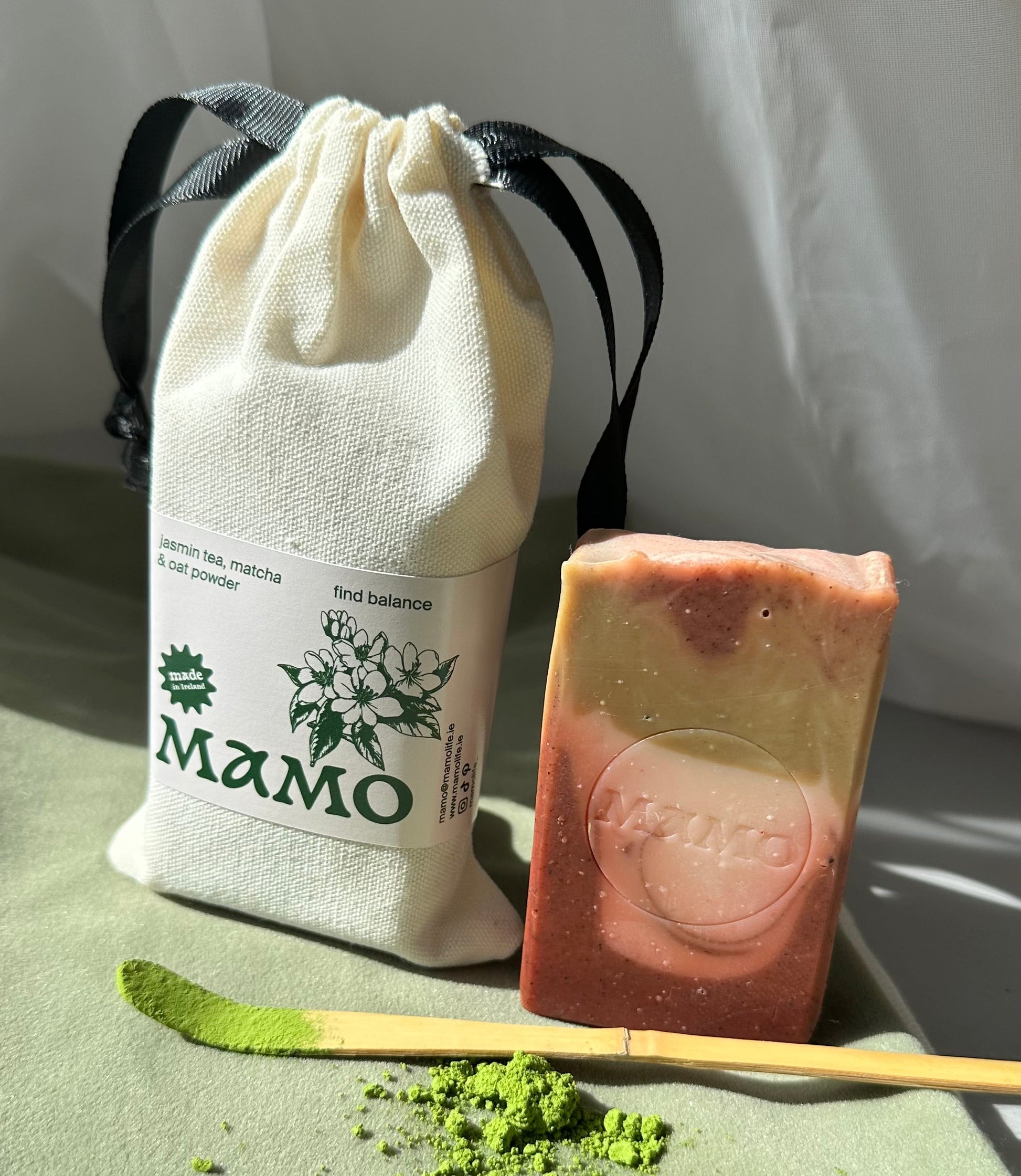 A handmade layered soap bar with pink, yellow, and green tones sits next to a cream-colored drawstring pouch with a black ribbon and a white label reading 'MAMO'. A bamboo spatula and green powder are also displayed on a soft green surface.