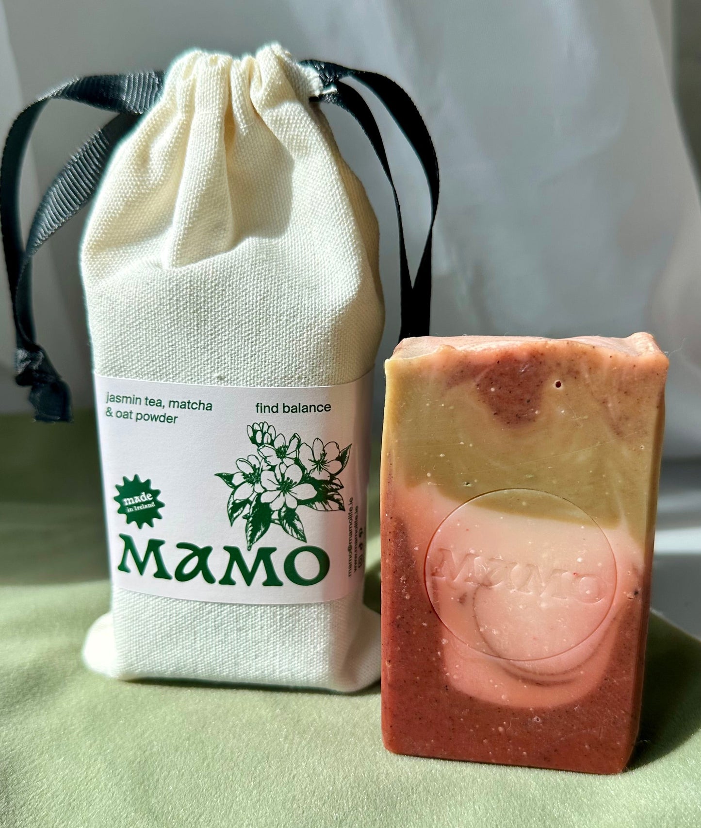 A handmade soap bar with pink and pale yellow marbled layers sits next to a small cream-colored drawstring pouch labeled 'MAMO' on a light green fabric surface.