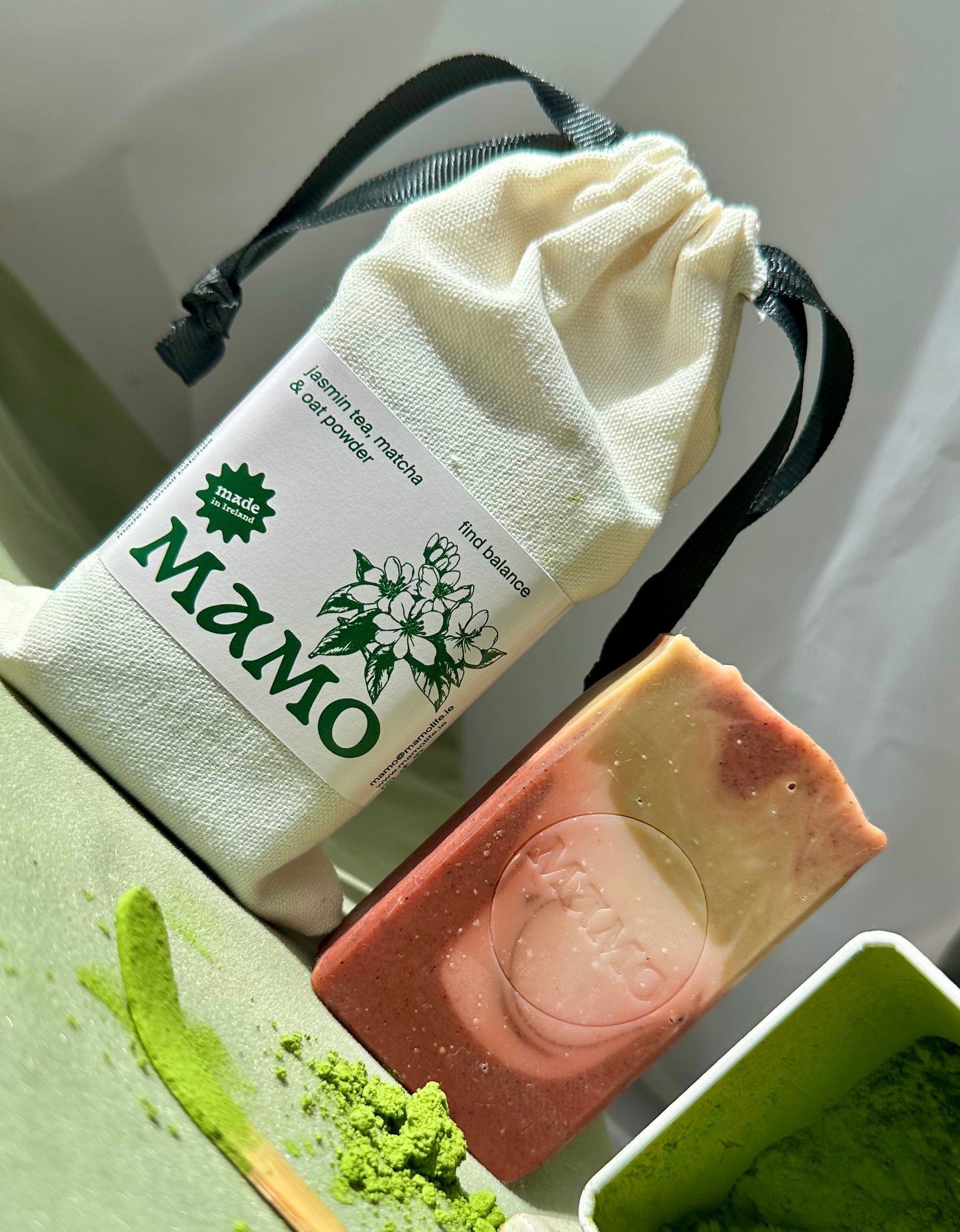 A handmade soap bar with pink and cream layers, displayed next to a white drawstring pouch labeled 'MAMO' and a green powder container on a light green surface.