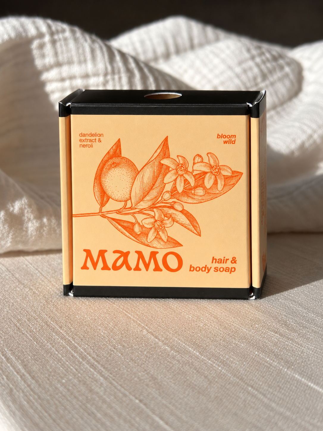 A rectangular box of MAMO hair & body soap sits on a textured beige fabric surface with soft white fabric in the background. The box is black with a peach-colored front label featuring orange botanical illustrations and the brand name MAMO.