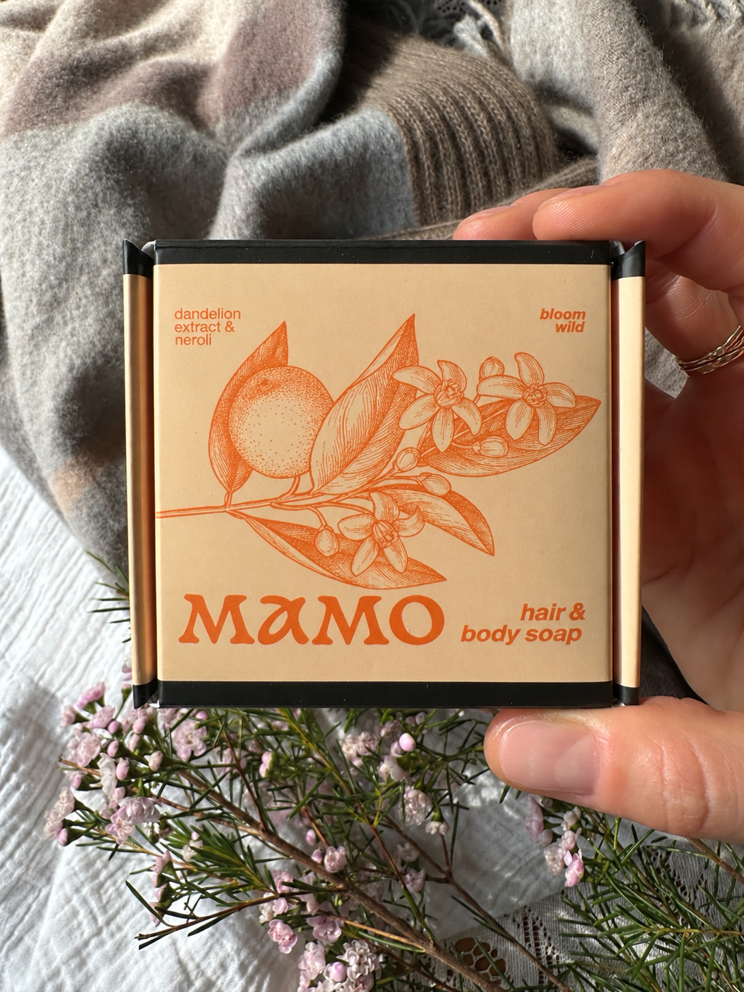 A hand holds a square box of MAMO hair and body soap. The box has a beige label with an orange botanical illustration of a citrus fruit and leaves, and the brand name 'MAMO' in large orange letters.