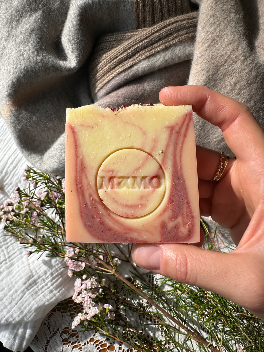 A hand holds a square handmade soap bar with a marbled cream and dusty rose pattern. The soap is embossed with the brand name 'MAMO' in the center.