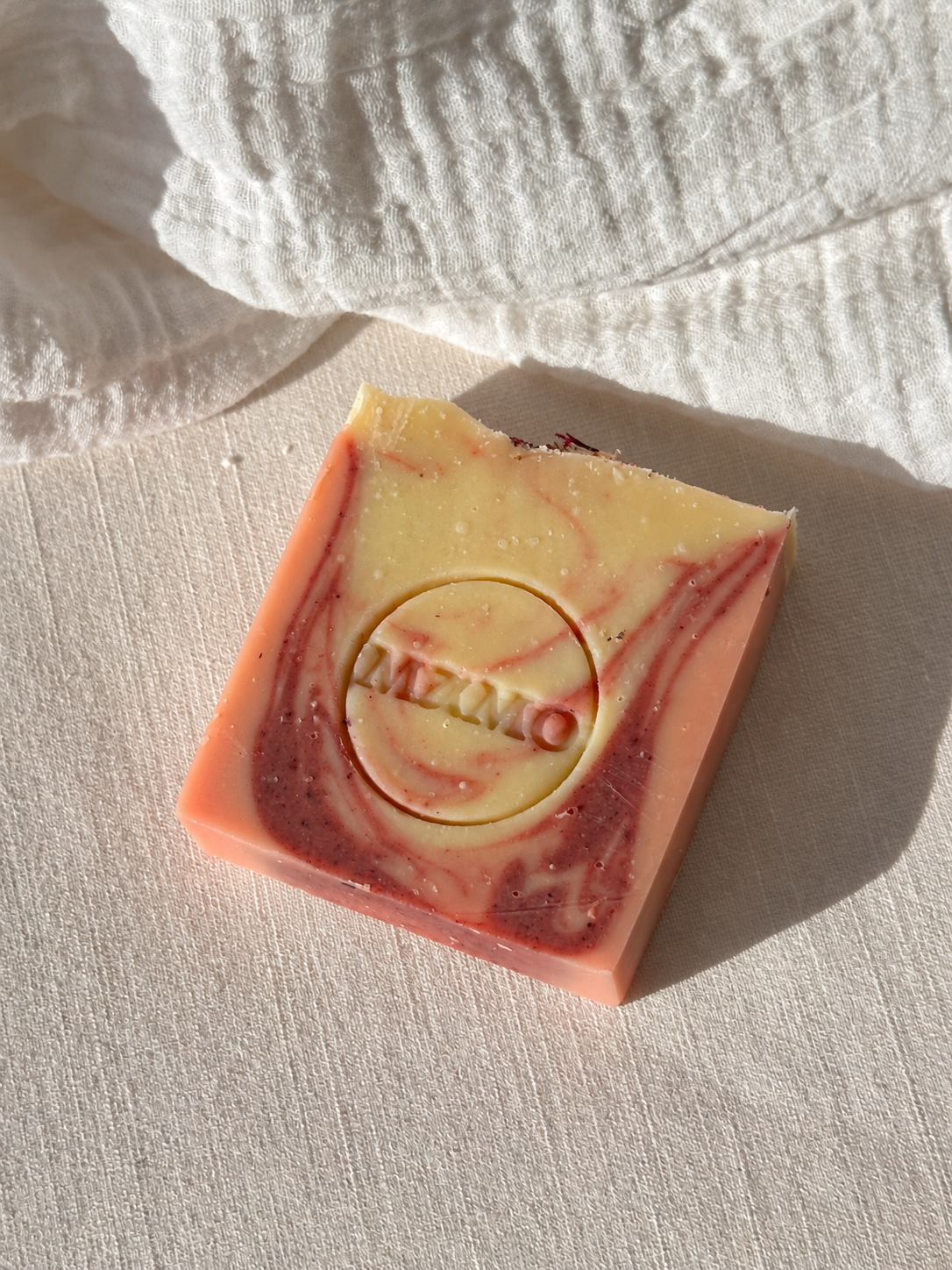 A rectangular bar of handmade soap with a marbled swirl pattern of pink, cream, and dark red, featuring a circular inset with the brand name 'MAMO' embossed. The soap is photographed on a textured off-white fabric under bright, direct sunlight.