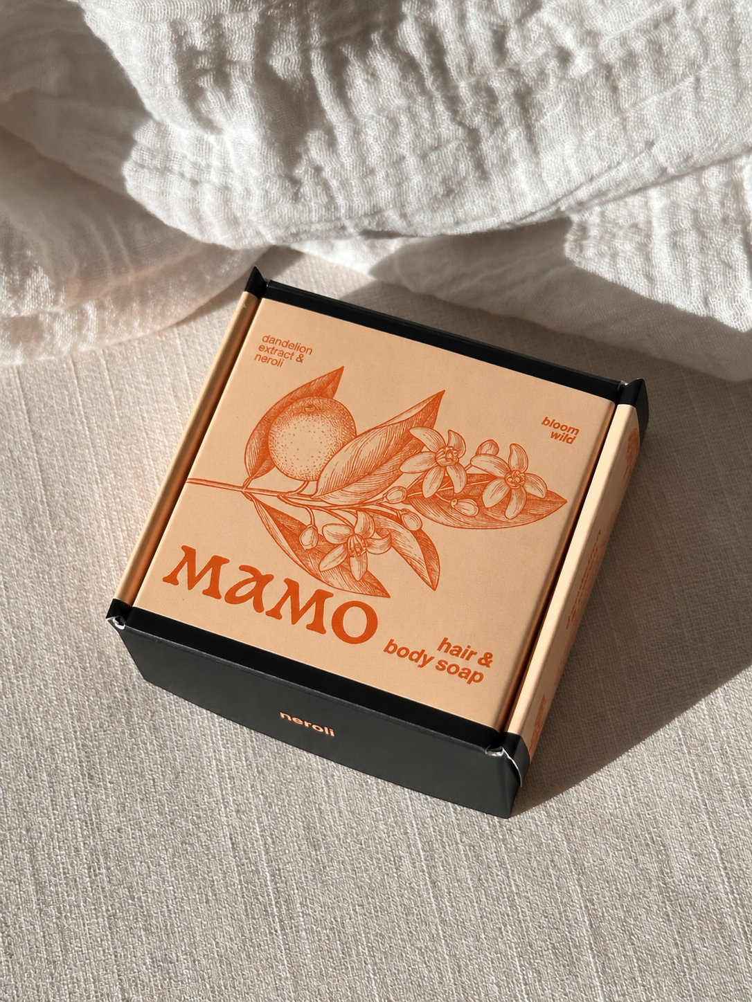 A boxed bar of MAMO hair & body soap placed on a textured cream fabric surface with soft natural shadows. The box is peach-toned with an orange botanical illustration and the brand name MAMO prominently printed, while the lower tray is black.