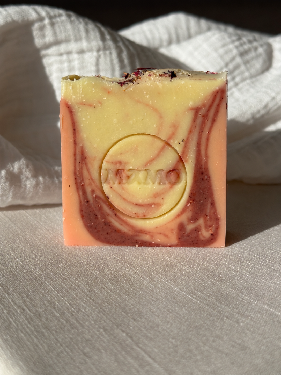 A rectangular handmade soap bar with a marbled swirl pattern of pale yellow, peach, and dark red, featuring a circular embossed logo in the center that reads 'MIXMO'. The soap is photographed standing upright on a textured white fabric surface with a matching fabric backdrop under direct sunlight.